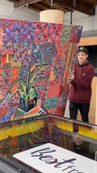 A large, brightly patterned painting lowered face down onto the glass of the flatbed scanner, a Best Framing team member holding its far edge