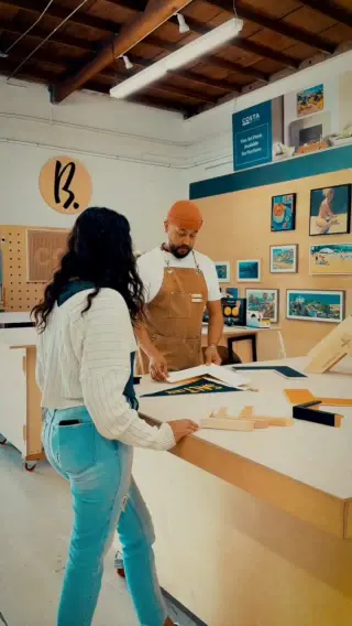A customer at the Best Framing design counter with a framer in a tan apron and orange beanie, framed prints and the painted B. roundel on the wall behind