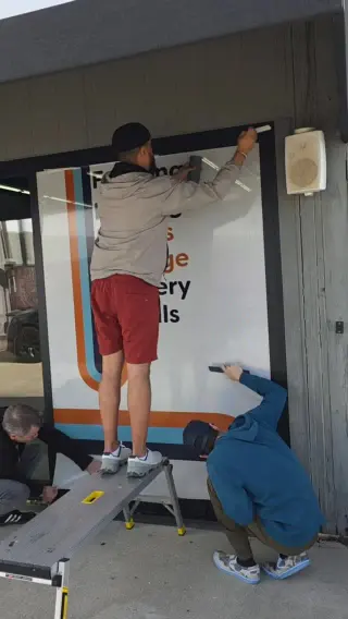 Three of the team fitting perforated vinyl to the shop’s own storefront window, one on a step ladder and two crouched at the sill