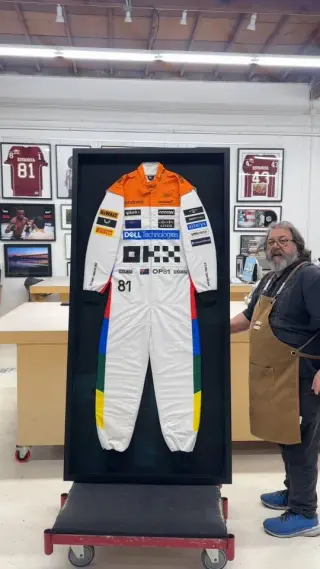 A race suit mounted at full length in a tall black shadow box on a cart, a framer in a tan apron beside it and framed jerseys on the wall behind