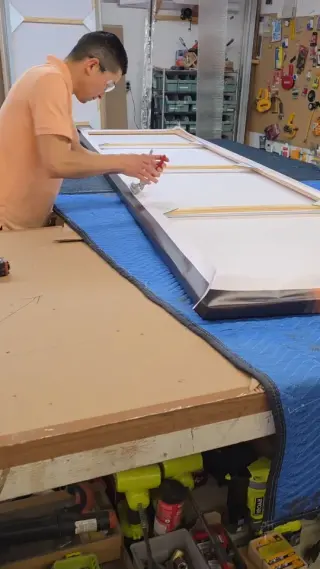 A framer working along a long floater frame laid out on a padded bench in the wood shop