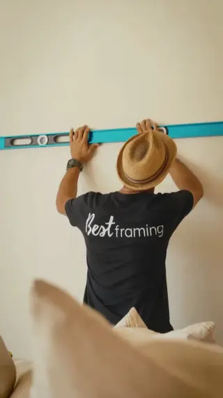 An installer in a Best Framing tee holding a long level against a bare wall above a sofa