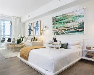 A white bedroom suite with a green and gold abstract in a slim white frame over the bed and gold-framed aerial photographs by the sofa