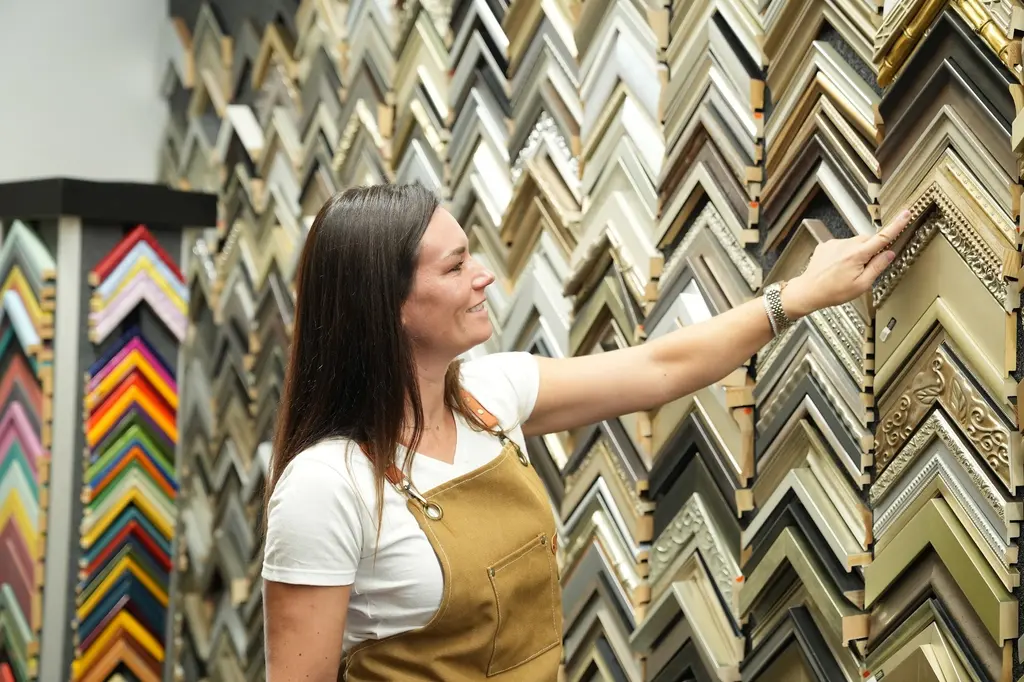 A Best Framing designer smiling as she reaches for a corner sample high on the moulding wall, a rack of colored corner samples beside her
