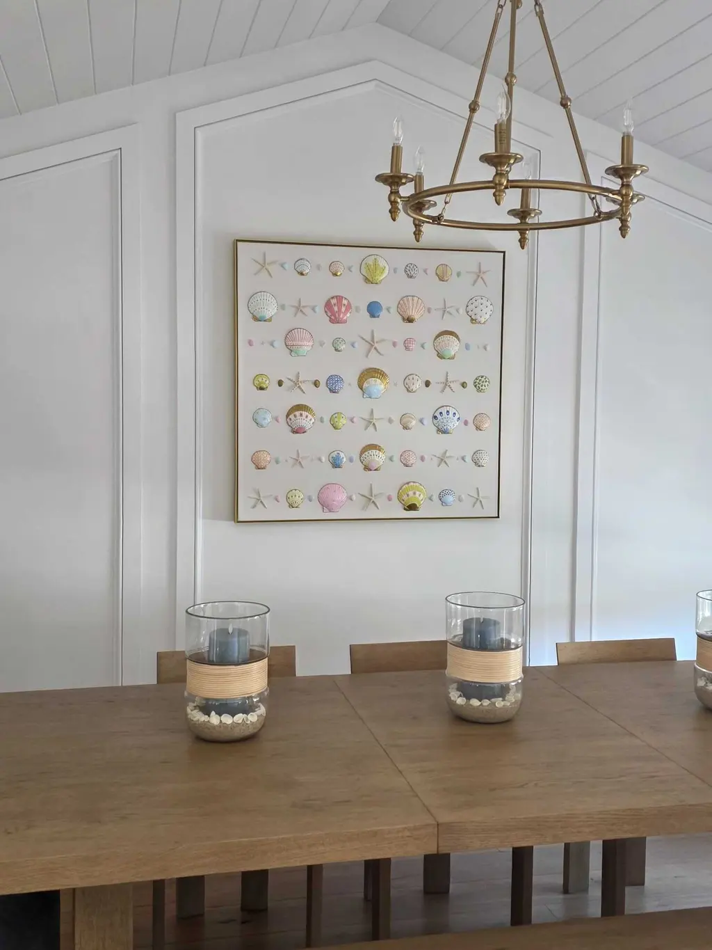 A framed grid of specimens over a farmhouse dining table under a brass chandelier