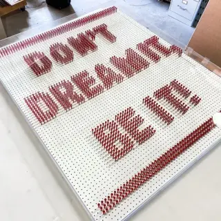 DON'T DREAM IT BE IT spelled in hundreds of red pegboard nails, in a clear acrylic box frame