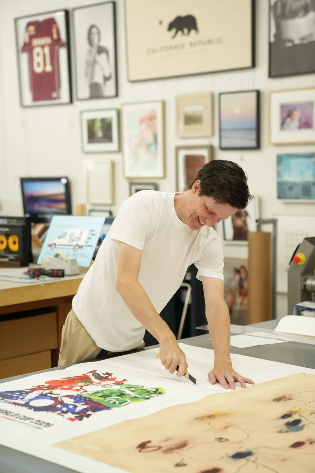 Mathew trimming the margin of a large print on the print-room table, framed work hung on the wall behind him