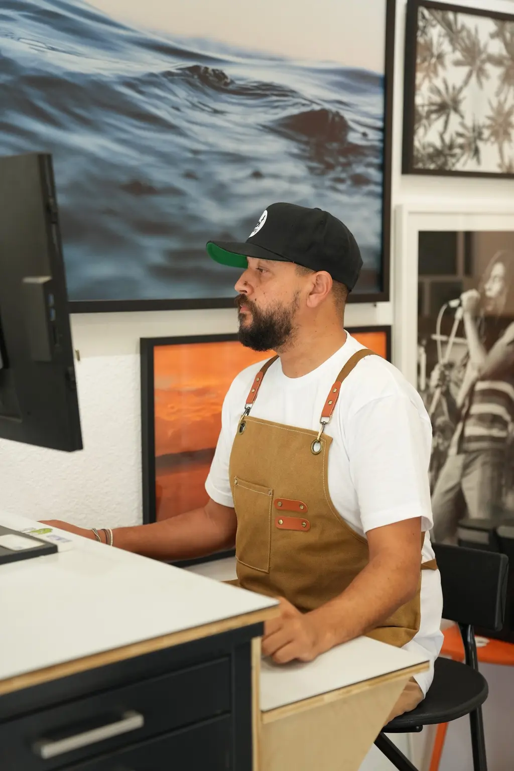 Medi at the print-lab computer, a large ocean photograph framed on the wall behind him