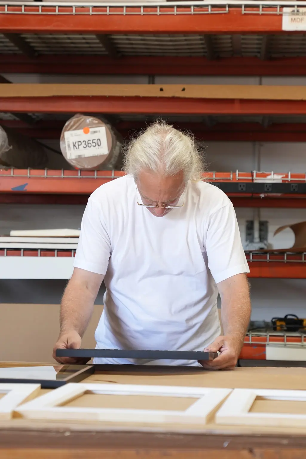 A framer with long white hair checking a black frame at the bench, cut white mats laid out in front of him and stock on the red shelves behind