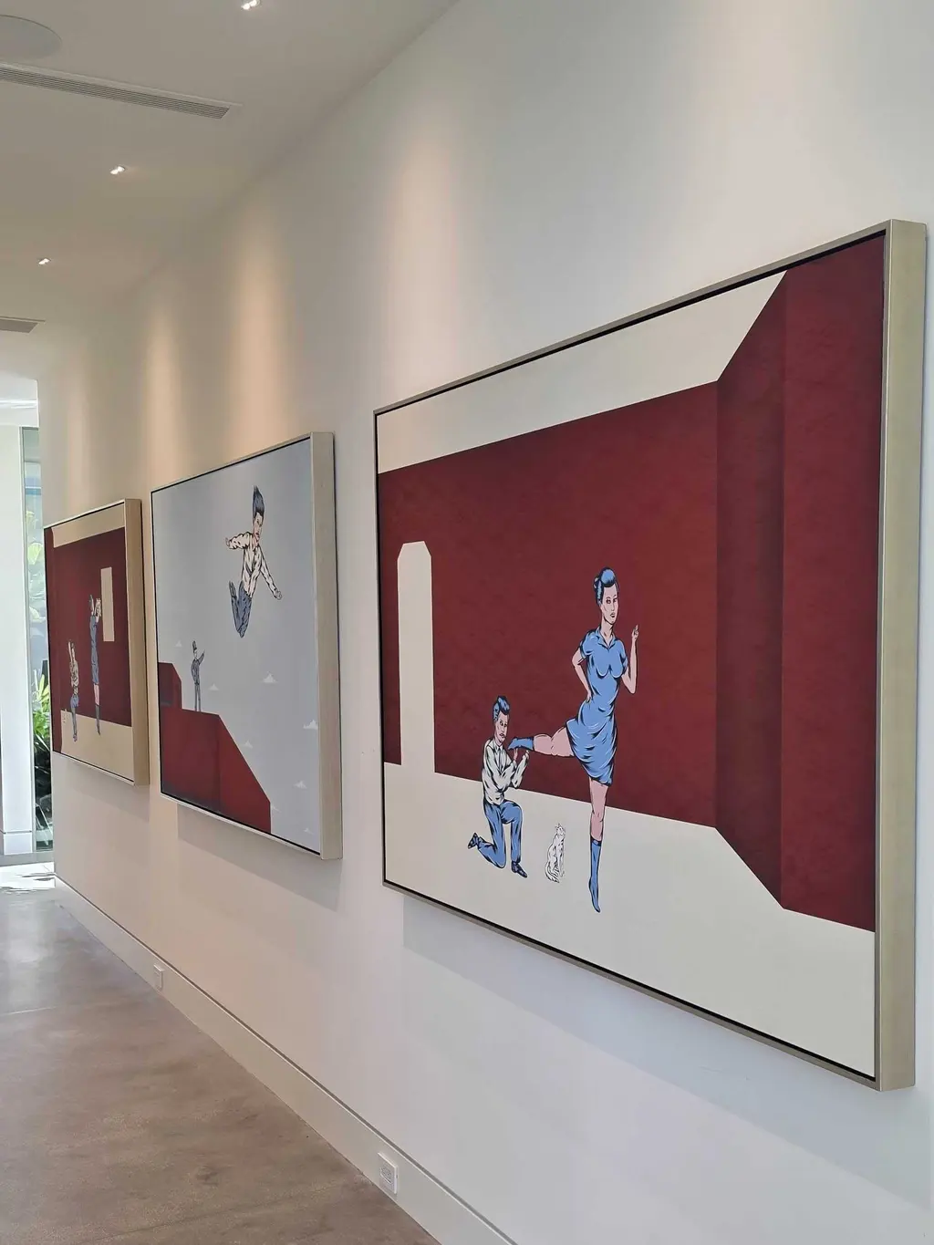 Three large canvases in pale floater frames hung in a row down a long white hallway, illustrated figures on deep red and grey grounds