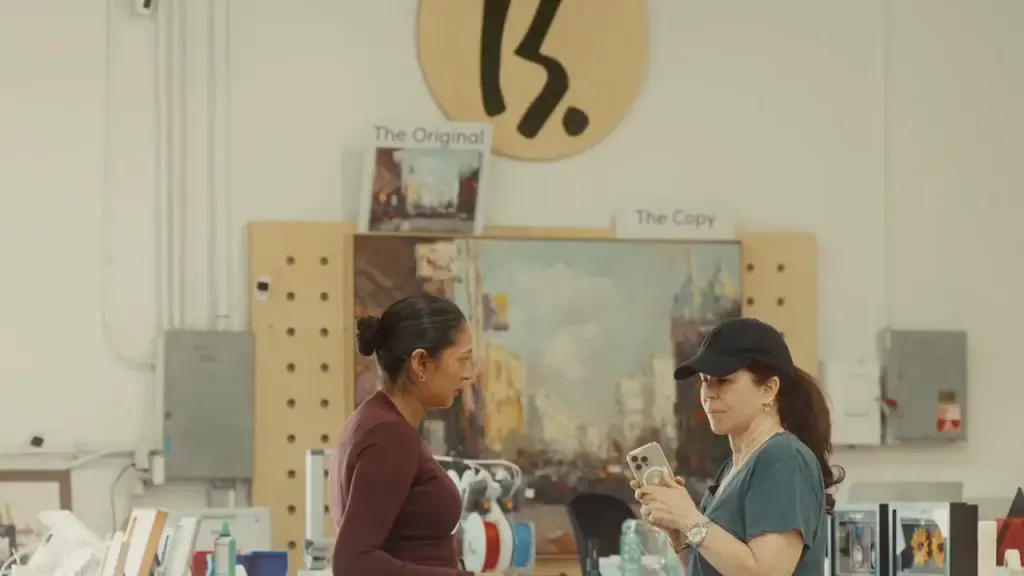 A small original street painting labelled The Original hung above a much larger copy labelled The Copy, two people talking at the counter in front of them