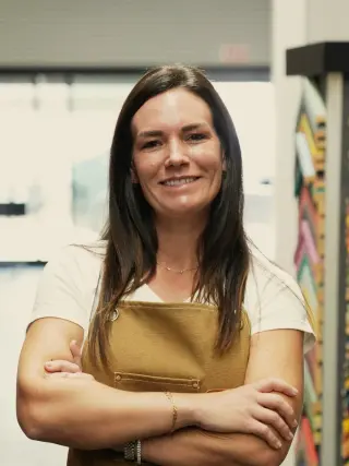Kristen in a canvas apron, arms crossed, beside a rack of mat samples
