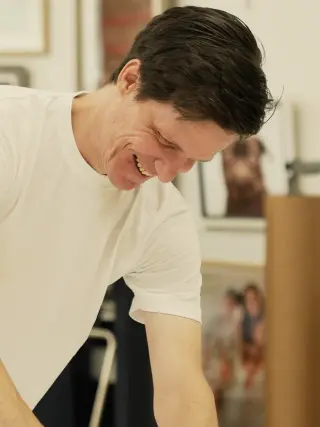 Mathew leaning over a print on the print-room table, framed work on the wall behind him