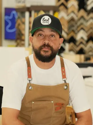 Medi in a Best Framing cap and canvas apron, seated in the showroom