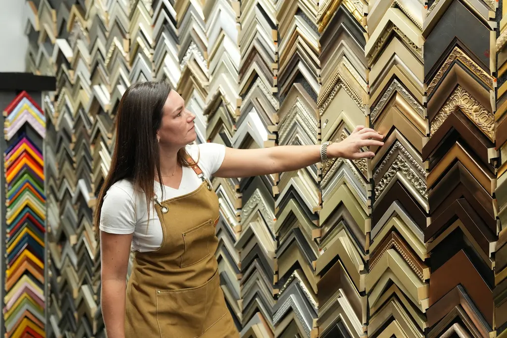 A designer reaching across the wall of moulding corner samples, carved gilt profiles under her hand