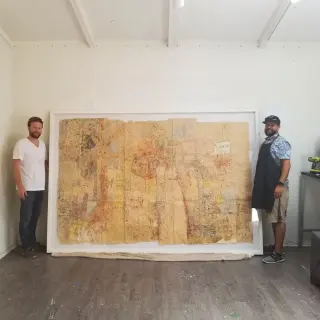 Huge ochre mixed-media map work in a white float frame, a framer standing at each edge for scale
