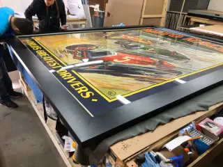 A vintage Auto Races World’s Greatest Drivers poster in a deep black frame, laid flat on the workshop bench mid-build