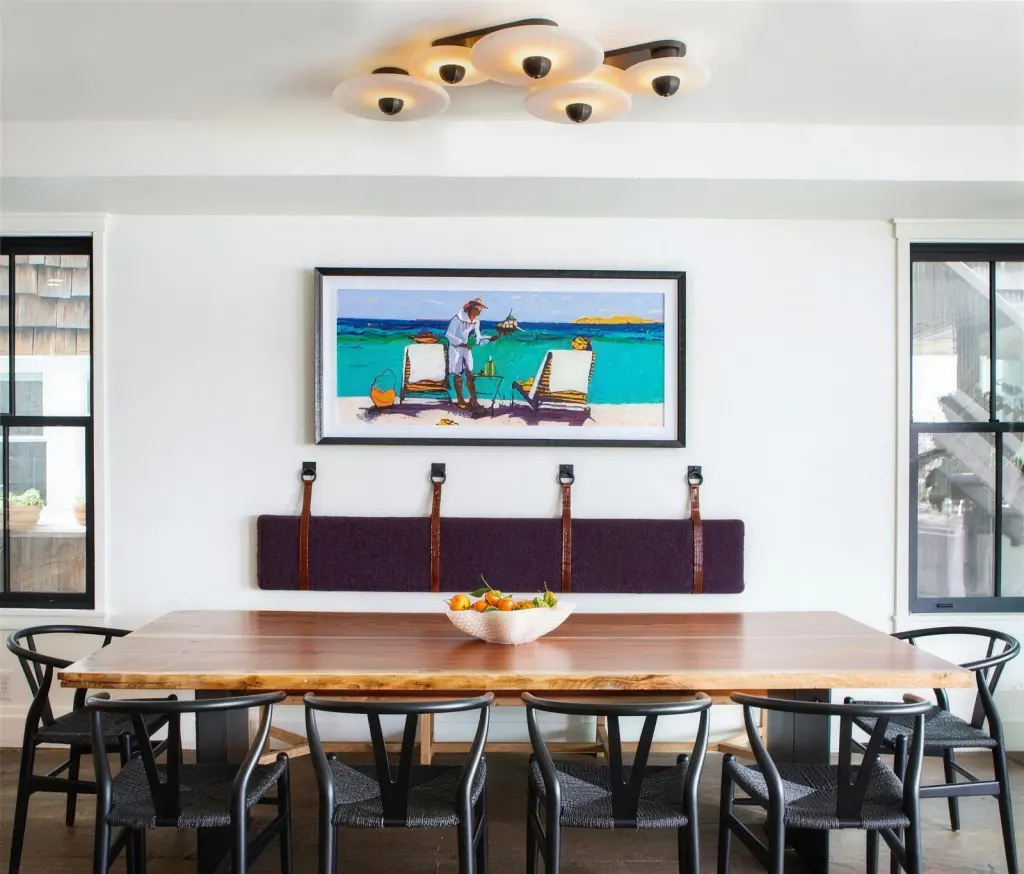 A framed beach painting in a slim dark frame above a leather-strapped banquette cushion and a live-edge dining table
