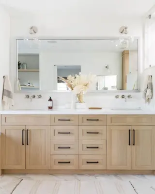One wide, pale-framed mirror spanning a white-oak double vanity with black pulls, dried flowers on the counter
