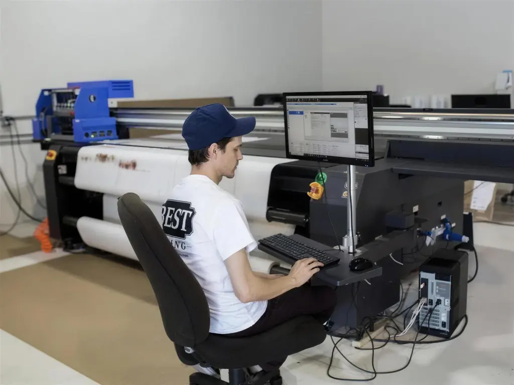 A technician at the print-lab workstation preparing a file on screen, the wide-format printer and racked rolls of media behind him