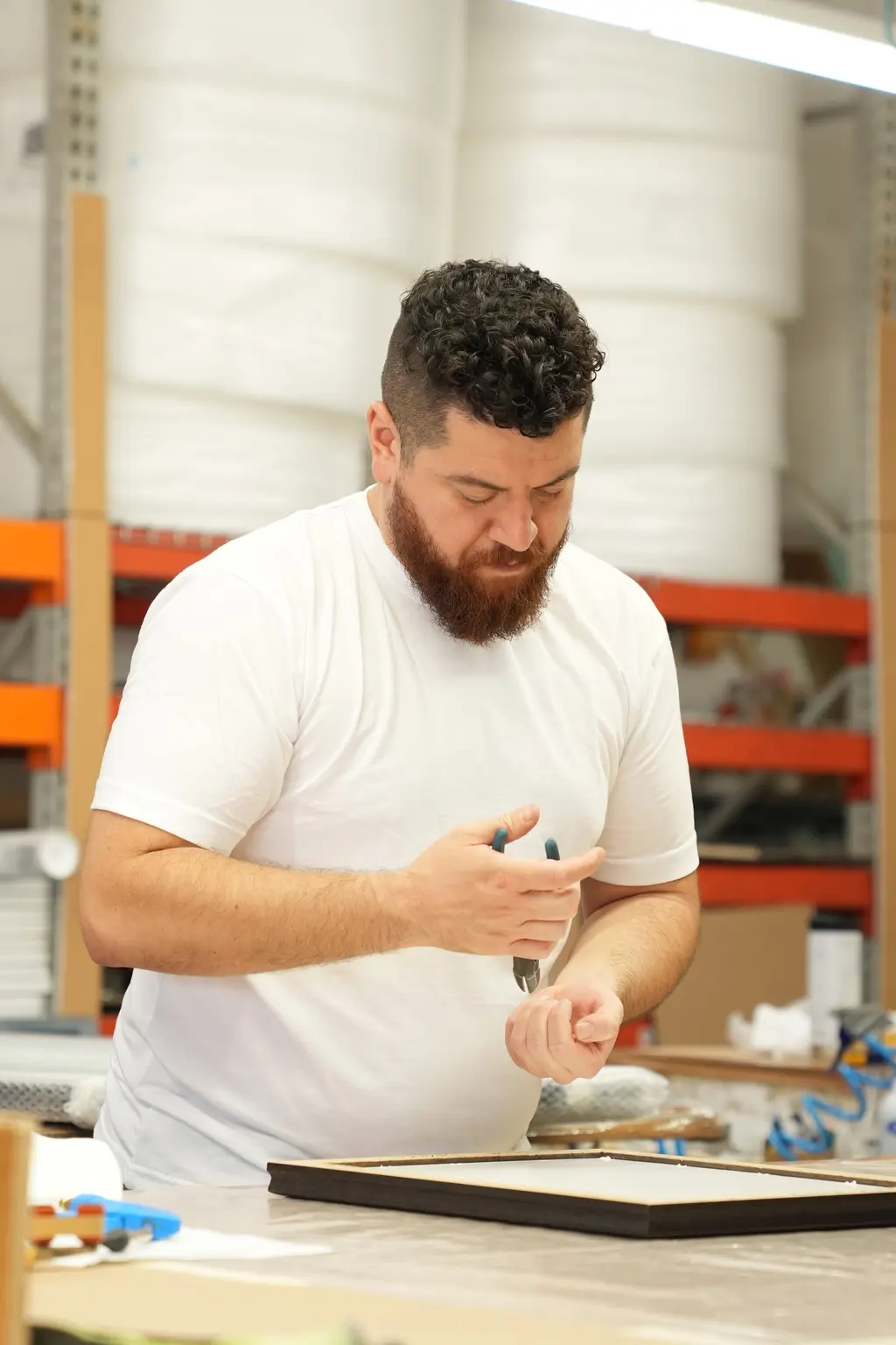 A framer with a red beard working a hand tool at the bench beside a black frame, rolls of bubble wrap on the shelves behind him