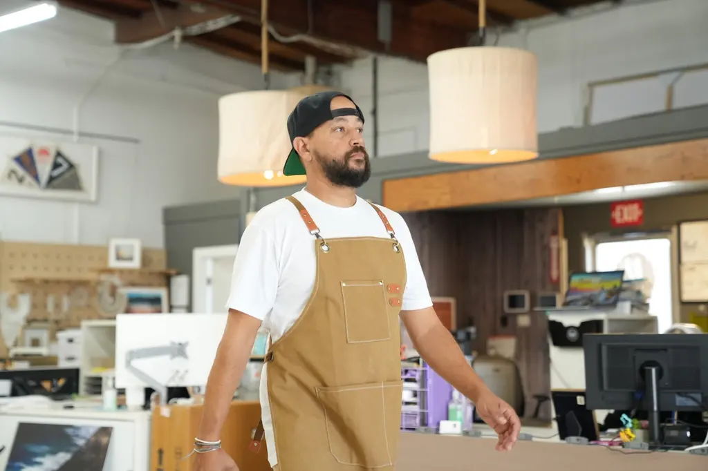 A team member in a tan canvas apron walking through the print lab, framed work and desks behind him