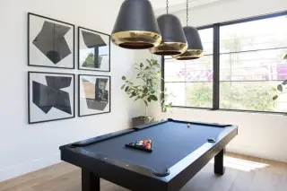 Four black-framed geometric abstract prints hung as a two by two set beside a navy felt pool table, three brass-lined pendants above it