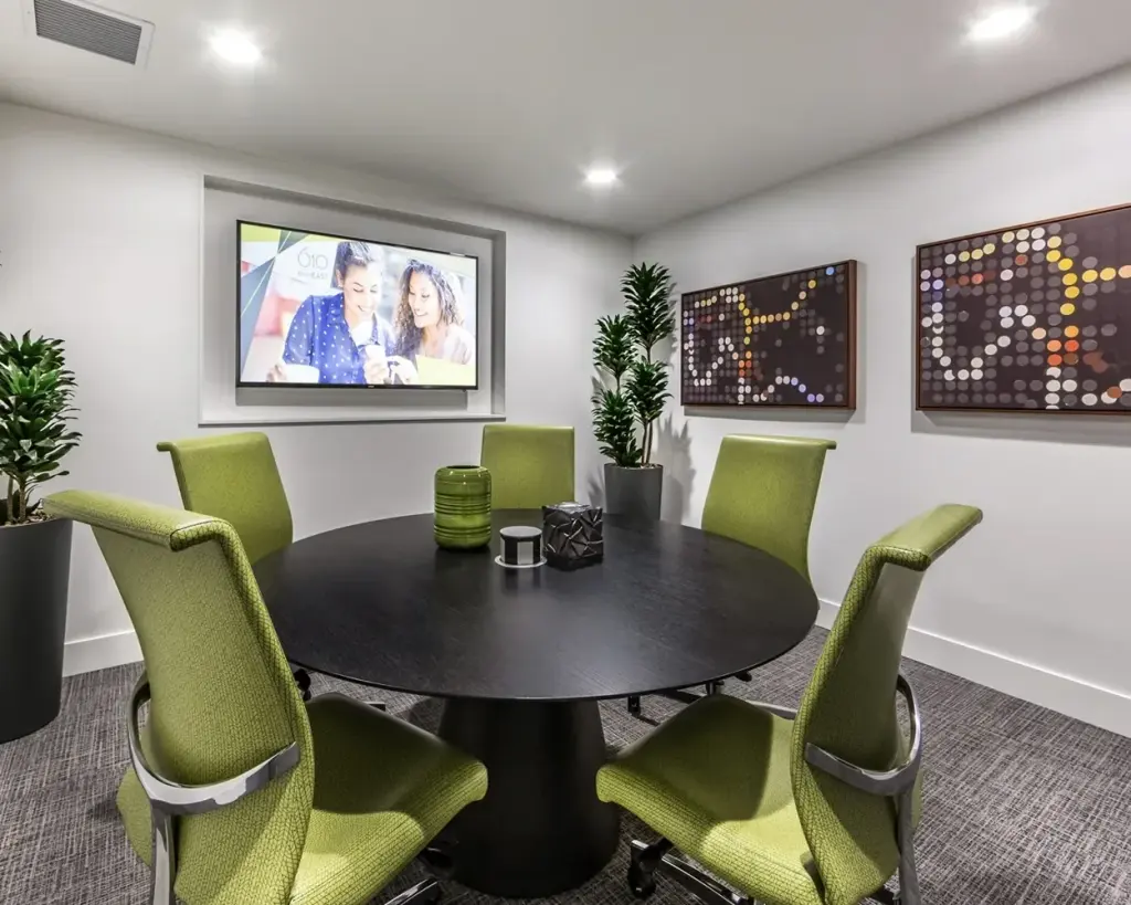 A small conference room with green chairs around a black round table, two framed dot-pattern artworks in wood frames beside the screen