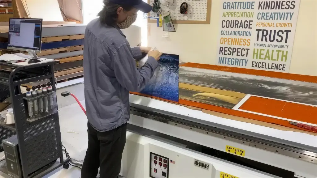 A technician in a cap and face mask lifting a printed ocean-wave panel from the bed of the flatbed printer, a long wave print still laid out beside it