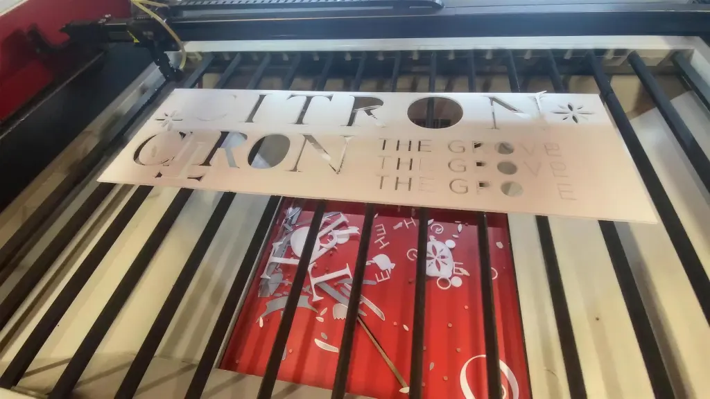 A cut sign panel on the laser bed, the lettering lifted out of a pale sheet with the material below showing through where the letters were