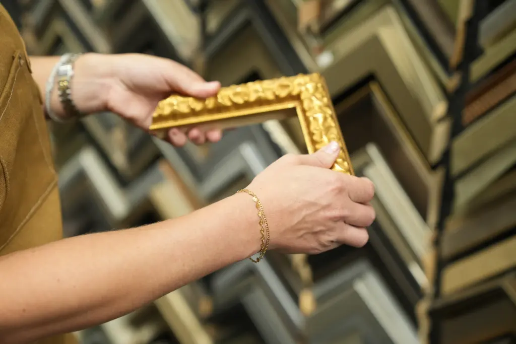 A hand holding a carved gilt corner sample in front of the wall of moulding corner samples