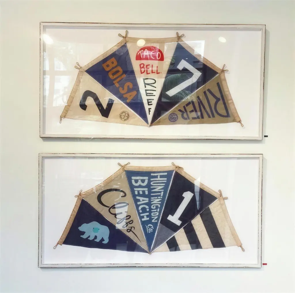 Two long white frames, one above the other, each holding a fan of vintage surf pennants that read Bolsa, River, Cliffs and Huntington Beach